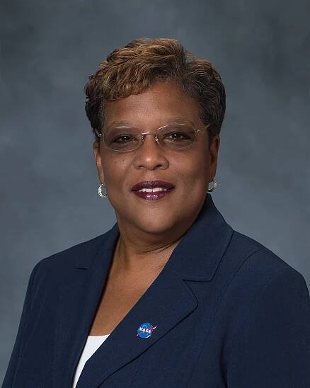 Portrait of a woman wearing glasses, pearl earrings, and a navy blazer with a NASA pin, smiling in front of a gray background.