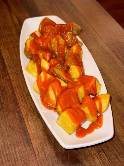 A white oval dish of golden fried potatoes drizzled with bright red spicy sauce, placed on a wooden table.