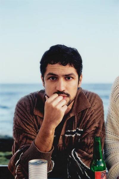 Gabriel Leone wears a brown sweater and sits with a green glass bottle on a beach.