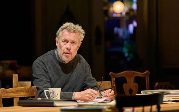 Alex Jennings sits on a wooden chair at a wooden table. He wears a grey jumper and holds a pair of glasses