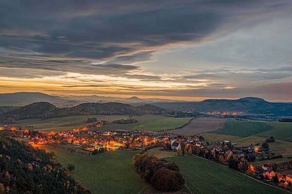 Ein malerisches Dorf unter einem dramatischen, bewölkten Himmel bei Sonnenuntergang. Die Landschaft ist geprägt von sanften Hügeln und vereinzelten Bäumen. Warmes Licht strahlt aus den Häusern und schafft eine gemütliche Atmosphäre. Am Horizont sind in der Ferne Berge zu sehen.