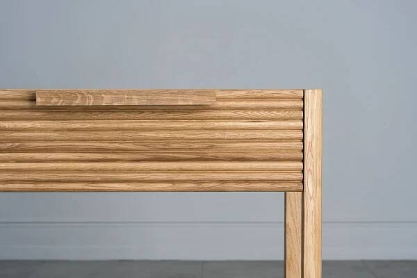 Tento odkládací stolek z masivního dubu kombinuje funkčnost s elegantním designem. Má dvě praktické zásuvky s charakteristickým drážkovaným čelem a minimalistickými úchyty, které dodávají moderní vzhled.