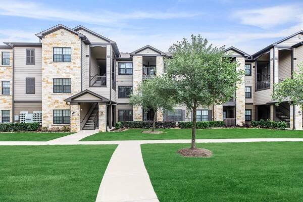 A modern apartment community featuring two to three-story buildings with a mix of stone and siding exteriors. The landscape includes well-maintained grassy areas, small trees, and paved walkways leading to the entrances. The sky is partly cloudy, creating a bright atmosphere.