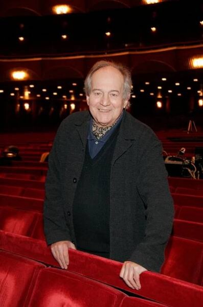 Otto Schenk, a man with thin white hair wearing an overcoat. stands in front of the orchestra seats in an empty opera house and smiles.