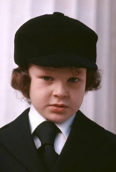 A child in formal attire with a black cap and tie stands facing the camera