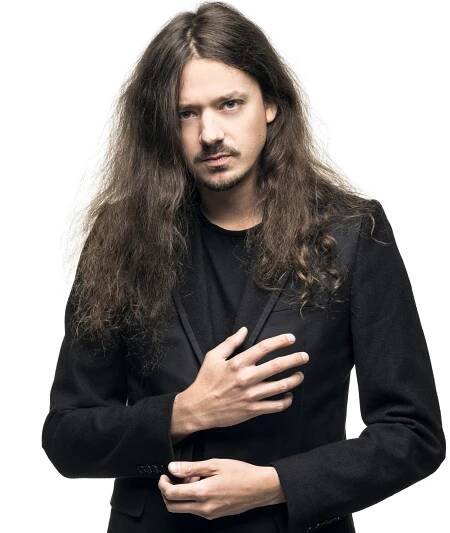 A man with a black shirt and a black jacket looks into the camera, with one hand on his chest and one hand on his stomach.