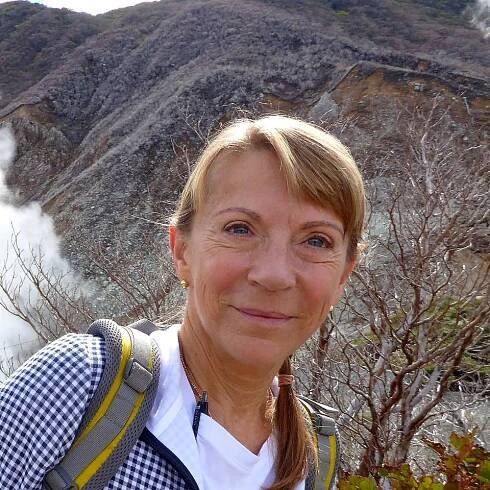 Frau vor einer dampfenden vulkanischen Landschaft mit kahlen Bäumen im Hintergrund.