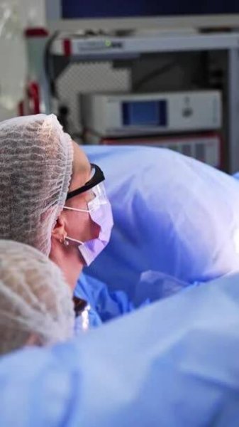 Dva lékaři s bílými čapkami a brýlemi sedí u pacientových nohou zakrytých modrým prostěradlem. Ženští chirurgové provádějí gynekologické operace. Svislé video. — Stock záběr
