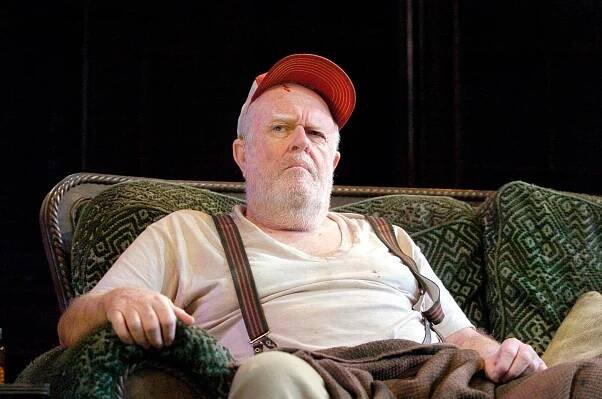 M. Emmet Walsh, with a white beard and wearing a light-colored T-shirt, overalls and a red baseball cap, sits on a couch onstage with an angry look on his face.
