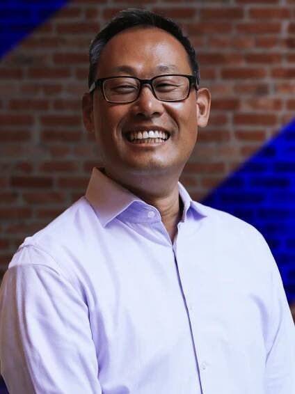 David Park, a smiling man in a light purple shirt standing in front of a brick wall with blue lights in the background.