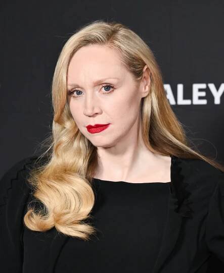 Person with long wavy hair, wearing an elegant black outfit, poses on a red carpet with a neutral expression