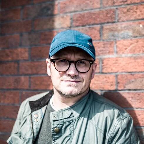 Lenny Abrahamson standing against a brick wall, wearing a blue cap, glasses, and a casual jacket.