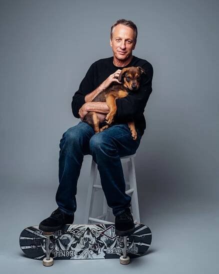 Man seated on a stool, wearing casual clothing, holding a small dog. A skateboard is placed below his feet