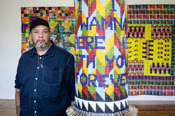 Jeffrey dressed in blue shirt stands next to colorful beaded artwork
