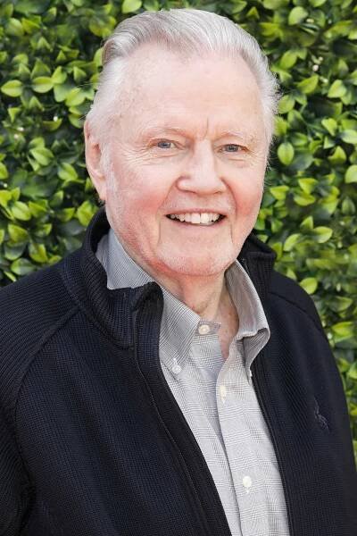 Jon Voight smiling, wearing a checkered shirt under a dark jacket, standing in front of a leafy backdrop