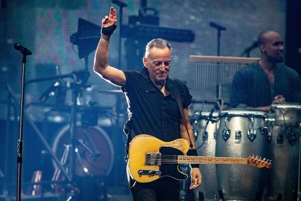 Bruce Springsteen onstage holding one hand in the air. He is wearing all black and has a yellow guitar around him.
