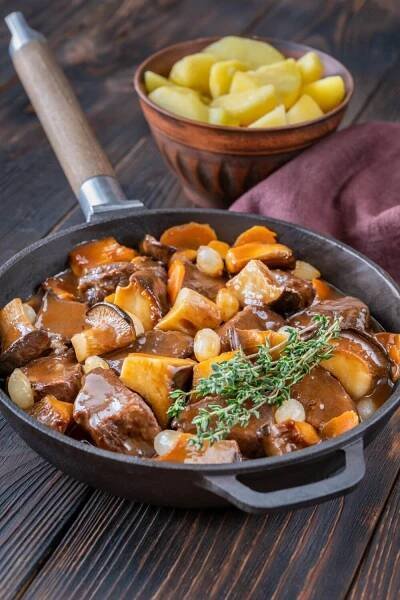 A hearty serving of Boeuf Bourguignon, a famous French beef stew, in a cast iron skillet, garnished with fresh thyme. The rich, savory sauce coats tender chunks of beef, carrots, mushrooms, and pearl onions. In the background, a bowl of boiled potatoes completes this traditional French meal, capturing the rustic and homey essence of Julia Child's classic recipe.