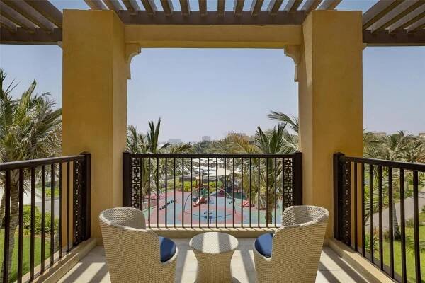 Doubletree By Hilton Resort & Spa Marjan Island Ras al-Khaimah Exterior photo The photo shows a balcony with two wicker chairs and a small round table, overlooking a resort pool area. The balcony is covered by a pergola with wooden beams and is painted a warm yellow color. The railing of the balcony is dark brown with decorati