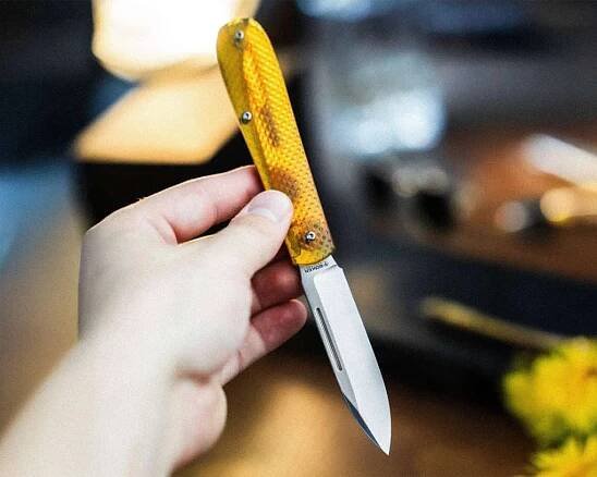 A hand holding an open folding knife with a yellow textured handle and a shiny silver blade. The background is blurred, showing indistinct objects and a hint of yellow flowers.