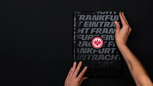 Eintracht Frankfurt Brandbook Cover. Zu sehen ist das Cover des Eintracht Frankfurt Brandbooks. Zwei Hände halten das Buch. Das schwarze Cover ist mit einer schwarzen Folienkaschierung veredelt. Die Veredelung zeigt den Eintracht Frankfurt Schriftzug. In der Mitte ist das Wappen des Vereins zu sehen.