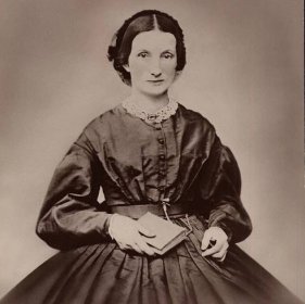 Black and White photograph of a women in a nineteenth-century dress holding a book in her right hand.