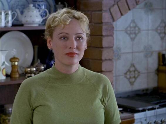 Virginia Madsen as Emma Burke in Returning Lily (AKA Tempted), wearing a light green sweater with a gentle, contemplative expression. She stands in a cozy, rustic kitchen with decorative plates and tiled walls in the background.