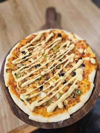 A fusion pizza, topped with chicken, green peppers, onions, and black olives, drizzled with a creamy sauce. It is placed on a wooden board. Spotted it at a restaurant in Kozhikode, Kerala. #WPNP2025