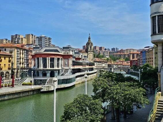 Bilbao's European cityscape features a river winding through traditional buildings, including a prominent market hall. A clock tower punctuates the skyline while trees grace the waterfront promenade. Modern apartments stand in the background, reminiscent of places to visit in Spain in 2025.