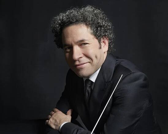 Gustavo Dudamel with curly hair in a suit leans against a dark table and holds a baton in front of a dark background. He has dimples and looks directly into the camera.