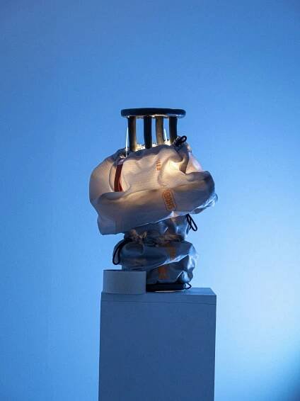 A shiny metal object in a white bag, illuminated against a blue backdrop, rests on a pedestal with tape beside it.