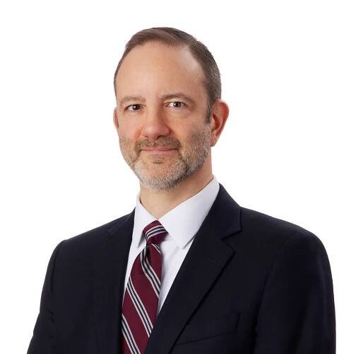 A man is dressed in a dark suit with a maroon, white, and gray striped tie against a plain white background.