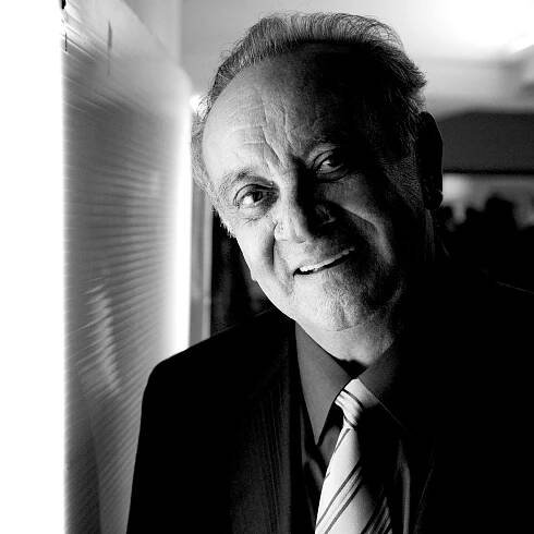 The late composer Angelo Badalamenti in a suit stands near a window smiling.
