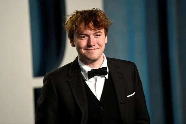 A young man in a tuxedo smiles on an arrival carpet.