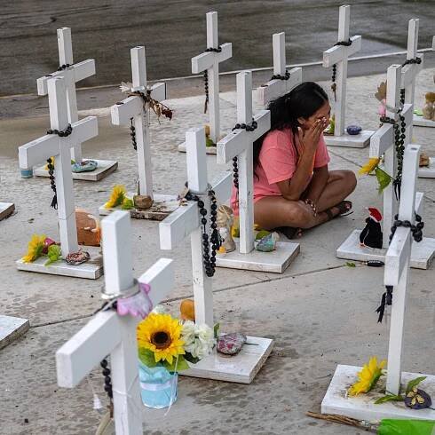 Ms. Rubio sits on a sidewalk cross-legged, in a pink T-shirt and sandals, wiping her eyes. She is in front of a small white cross, surrounded by other white crosses, decorated with sunflowers and rosaries.