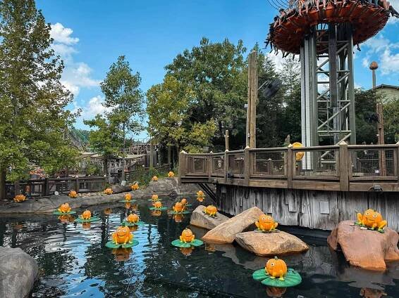 A round ride with people on it beside a pond of water with pumpkins made to look like frogs on Lilypad's. 