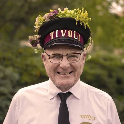 A Tivoli employee in uniform with a flower-adorned hat smiles warmly at the camera, with green trees in the background