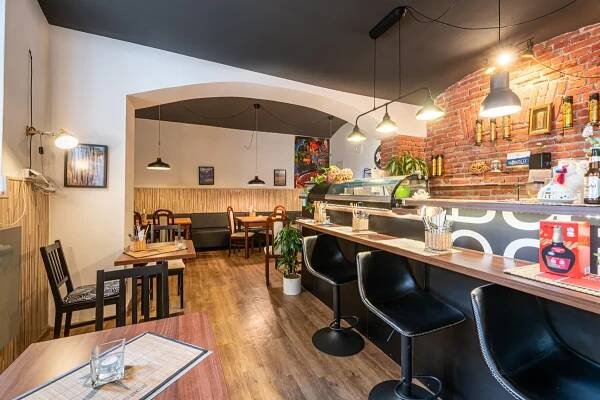 Interior view of a restaurant with wooden floors, tables, chairs, and a bar with black stools. A brick wall and hanging lights add to the ambiance.