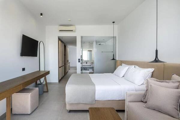 Michelangelo Resort & Spa Ágios Fokás Exterior photo The photo shows a modern and bright hotel room. The room is primarily white with light gray flooring. A large bed with a light beige headboard and white linens is the central feature, positioned against a white wall. Two pillows are placed on the bed