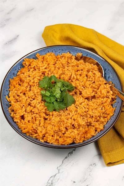 Red rice in a blue patterned serving bowl with spoon, garnished with coriander and with a mustard yellow napkin on the side