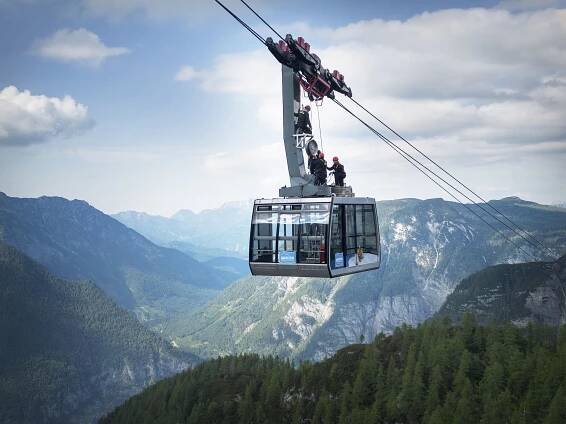 Seilbahnkabine mit Technikern auf dem Dach schwebt über grüne Hänge und Täler im Dachsteinmassiv.