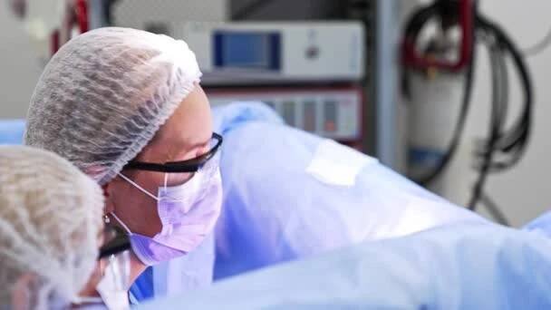 Main female doctor in cap, mask and protective glasses leans to the patient at surgery. Modern equipment in surgery room at blurred backdrop. — Stock Footage