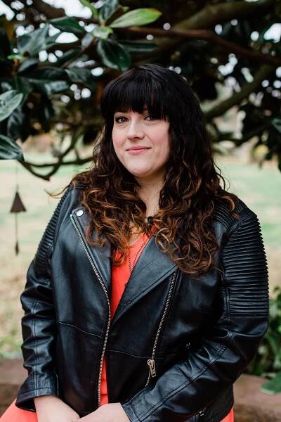 Woman smiling with shoulder length wavy hair, a black jack and a red shirt against a background of trees.