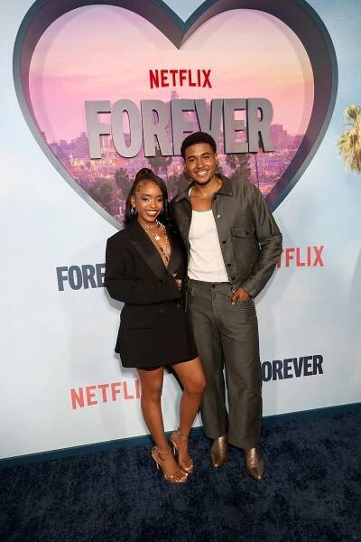 Two people on a Netflix event red carpet. One wears a chic blazer dress, and the other wears a casual jacket and pants. They smile at the camera