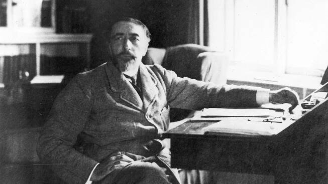 Der Schriftsteller Joseph Conrad (1857-1924) sitzt mit übergeschlagenen Beinen seitlich an einem Schreibtisch.