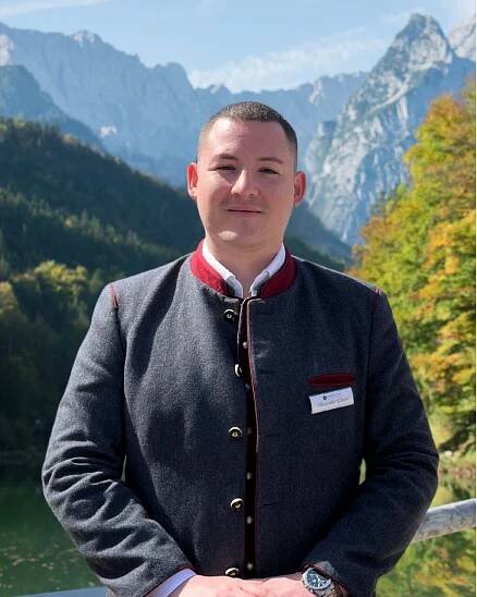 Portraitfoto von dem Restaurantleiter Alexander Glaser. Im Hintergrund sind die Berge und der Riessersee zu sehen.