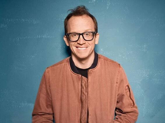 Chris Gethard wearing a brown jacket and glasses standing against a blue background.