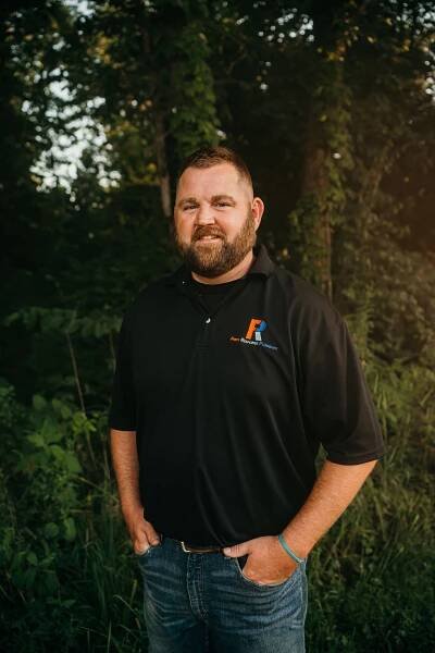 A man with a beard is standing in a field with his hands in his pockets.