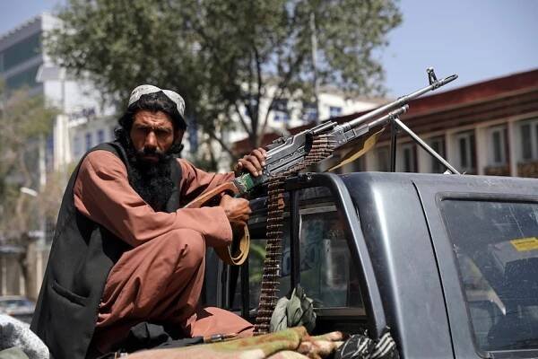 A bearded man with a giant rifle sits in the tray of a ute, looking at the camera 
