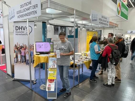 Das Bild zeigt die linke Seite des Messestands der Deutschen Gesellschaft Hörbehinderter auf der RehaCare 2024. An dieser Ecke steht das rollup des DHV und ein Monitor zeigt eine Endlospräsentation. Susanne Schmidt im DHV-T-Shirt (mit Aufdruck "HörExpertin") bietet das Faltblatt des DHV an.