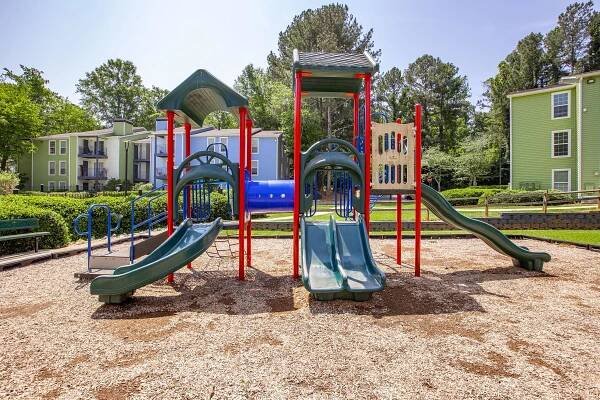 Playground featuring two green slides, climbing structures, and red support beams, surrounded by wood chips. In the background, there are green trees and residential buildings, creating a vibrant outdoor play area. Sunlight enhances the cheerful atmosphere. Perfect for children to enjoy.